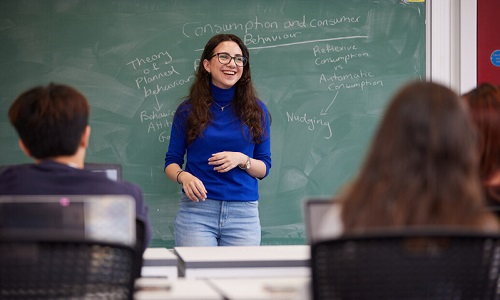 female student standing in a classroom front of green chalk board and smiling. Students are seated in the foreground facing her. 